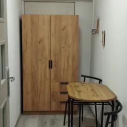 Ustawiony w rogu pokoju zestaw mebli: drewniana szafa z dwiema parami drzwi i trzema szufladami oraz niewielki stół z czterema krzesłami o metalowej konstrukcji i drewnianych siedziskach.