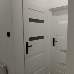 MAPKIM remonty - Dwie białe, nowoczesne drzwi wewnętrzne z czarnymi klamkami i poziomymi przeszkleniami, widoczne na tle jasnych ścian i podłogi ułożonej w jodełkę.