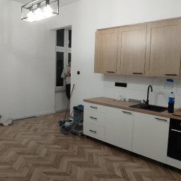 MAPKIM remonty - Nowoczesna kuchnia z białymi szafkami i drewnianym blatem, widok na parkiet w jodełkę, osoba w oknie i sprzęt budowlany w rogu pomieszczenia.