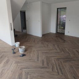 MAPKIM remonty - Ułożona podłoga z paneli w jodełkę w pustym, świeżo wyremontowanym pokoju z białymi ścianami; widoczne wiadro z gruntem Śnieżka, wkrętarka akumulatorowa i rękawice robocze.