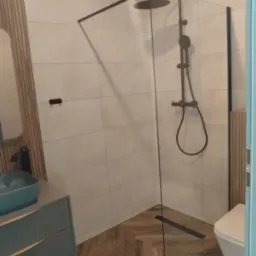 Nowoczesna łazienka z kabiną prysznicową typu walk-in z czarnym osprzętem, umywalką w kolorze turkusowym i drewnianą okładziną ściany.