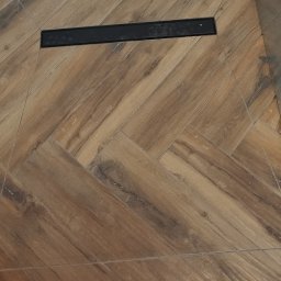 MAPKIM remonty - Ułożona w jodełkę podłoga z płytek imitujących drewno, z czarnym odpływem liniowym widocznym w górnej części kadru.