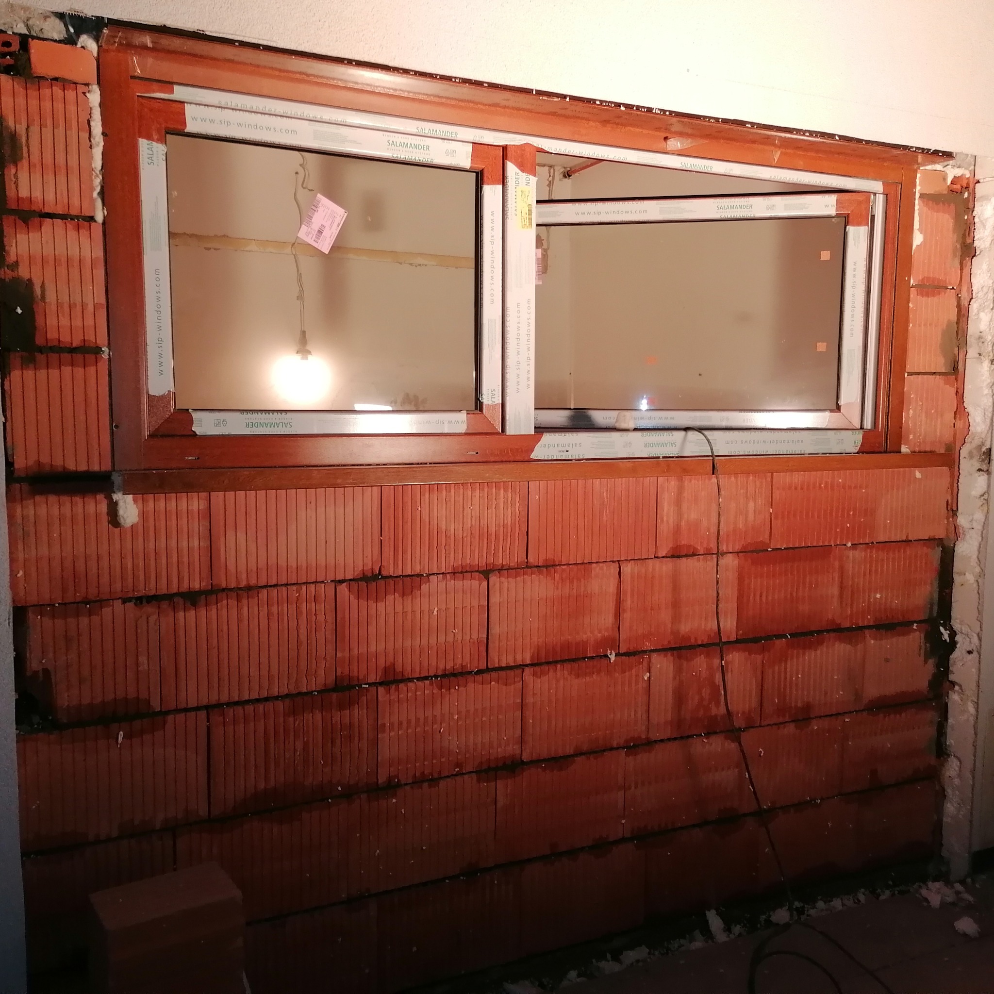 Montaż okna z brązową ramą marki Salamander w ścianie z czerwonej cegły ceramicznej dziurawki, widoczne szczeliny wypełnione pianką montażową i kabel zwisający ze ściany.