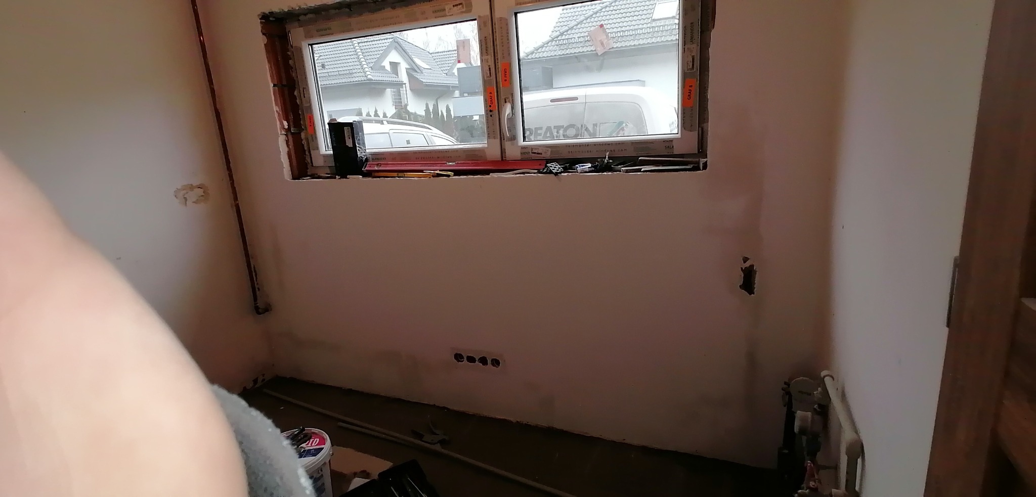 Wnętrze łazienki w trakcie remontu, widoczne okno z narzędziami na parapecie, ściany przygotowane do dalszych prac, puszki elektryczne i rury instalacji wodnej.