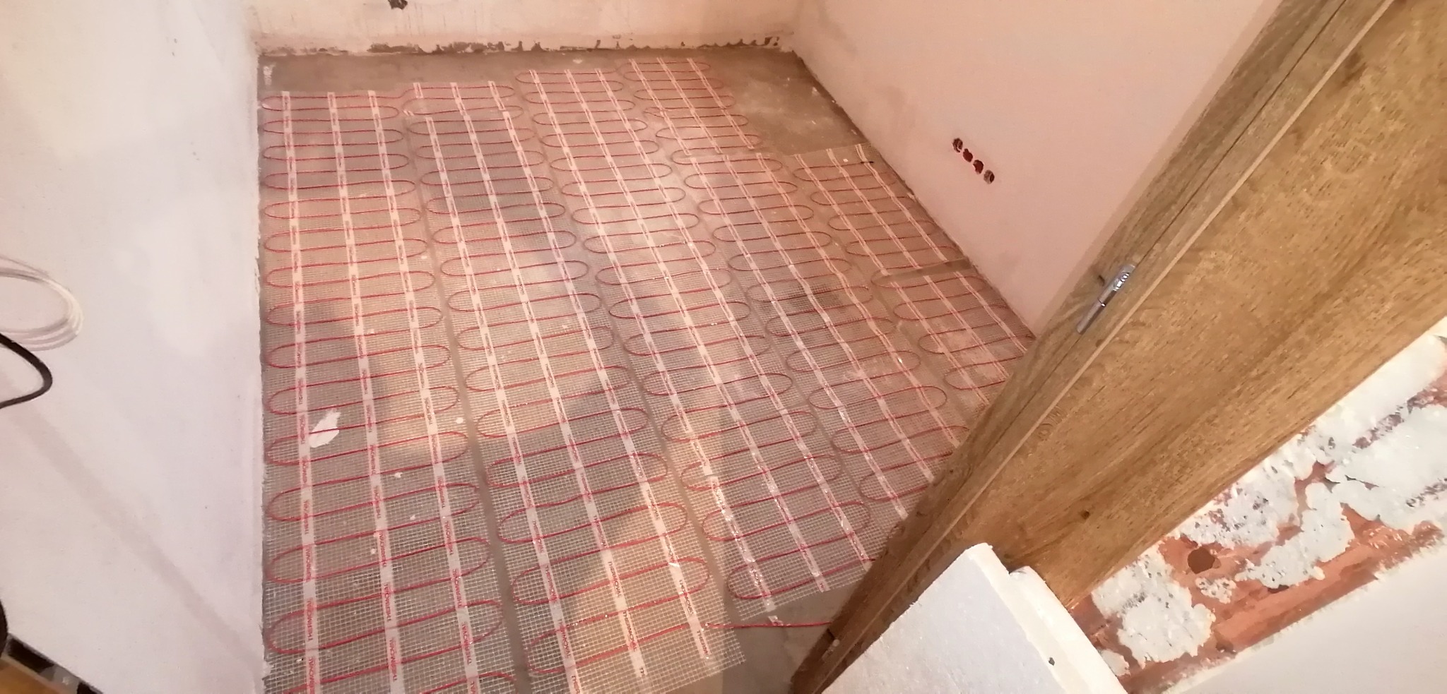 Rozłożona mata grzewcza na podłodze w trakcie remontu, widoczne przewody elektryczne, ściany w stanie surowym.