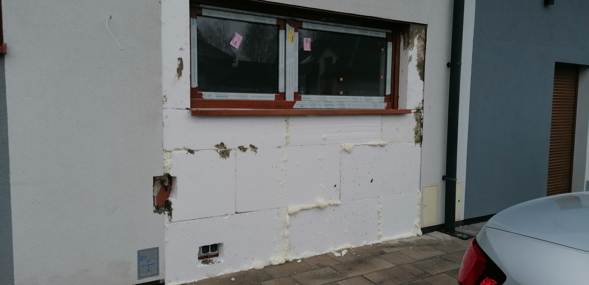 Ocieplanie ściany zewnętrznej budynku styropianem, widoczne okno z naklejkami, fragment samochodu i ściana sąsiedniego budynku.