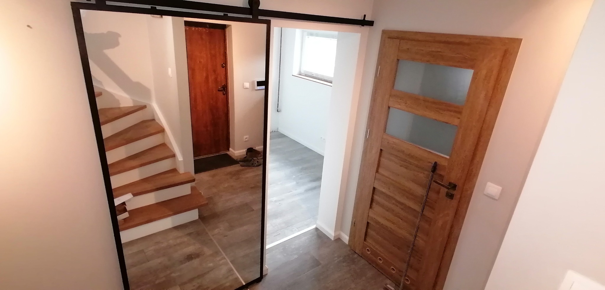 Wnętrze domu z przesuwnymi drzwiami lustrzanymi, odbijającymi schody i wejście, obok drewniane drzwi z przeszkleniami.