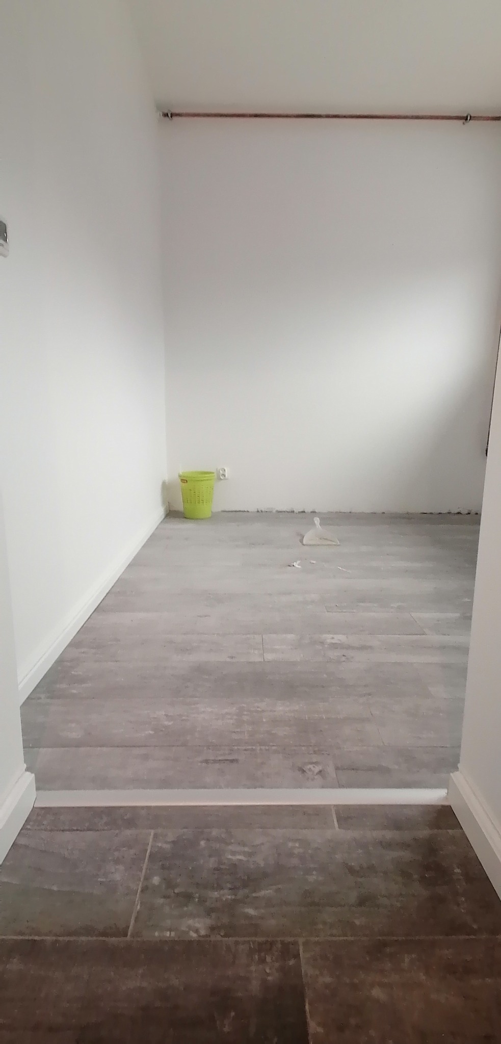 Wnętrze po remoncie z nową podłogą z płytek imitujących drewno, widoczny zielony kosz na śmieci i rurka miedziana pod sufitem.