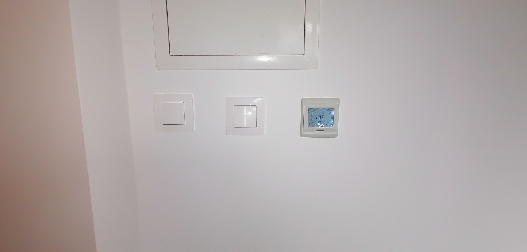 Biała ściana z klapą rewizyjną, pojedynczym i podwójnym włącznikiem światła oraz termostatem pokazującym temperaturę 20.5 stopni Celsjusza.