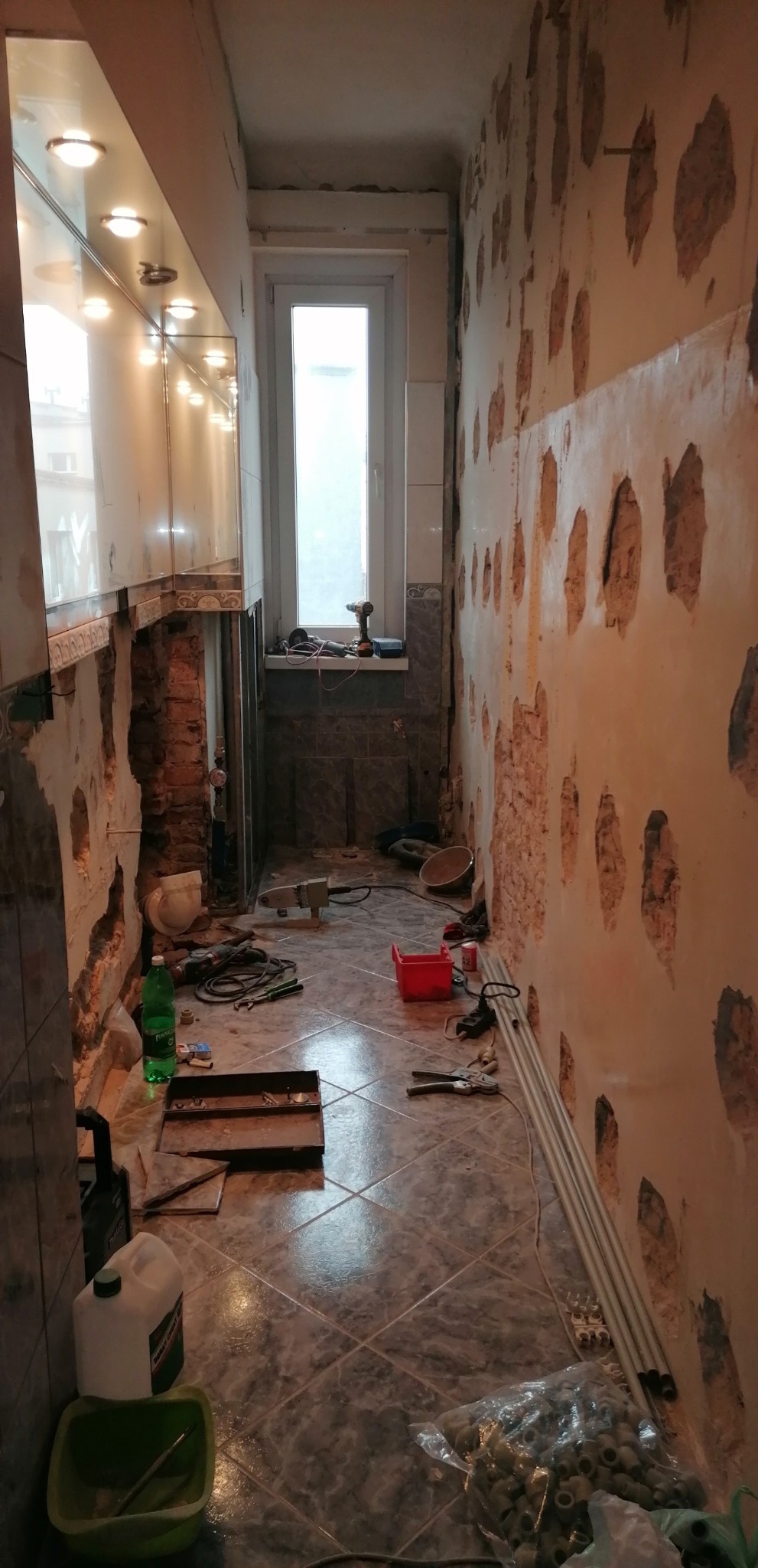 Wnętrze łazienki w trakcie remontu: ściany z odkrytą cegłą, częściowo położone płytki, narzędzia i materiały budowlane rozłożone na podłodze z szarych płytek, widoczne rury PCV i skrzynka...