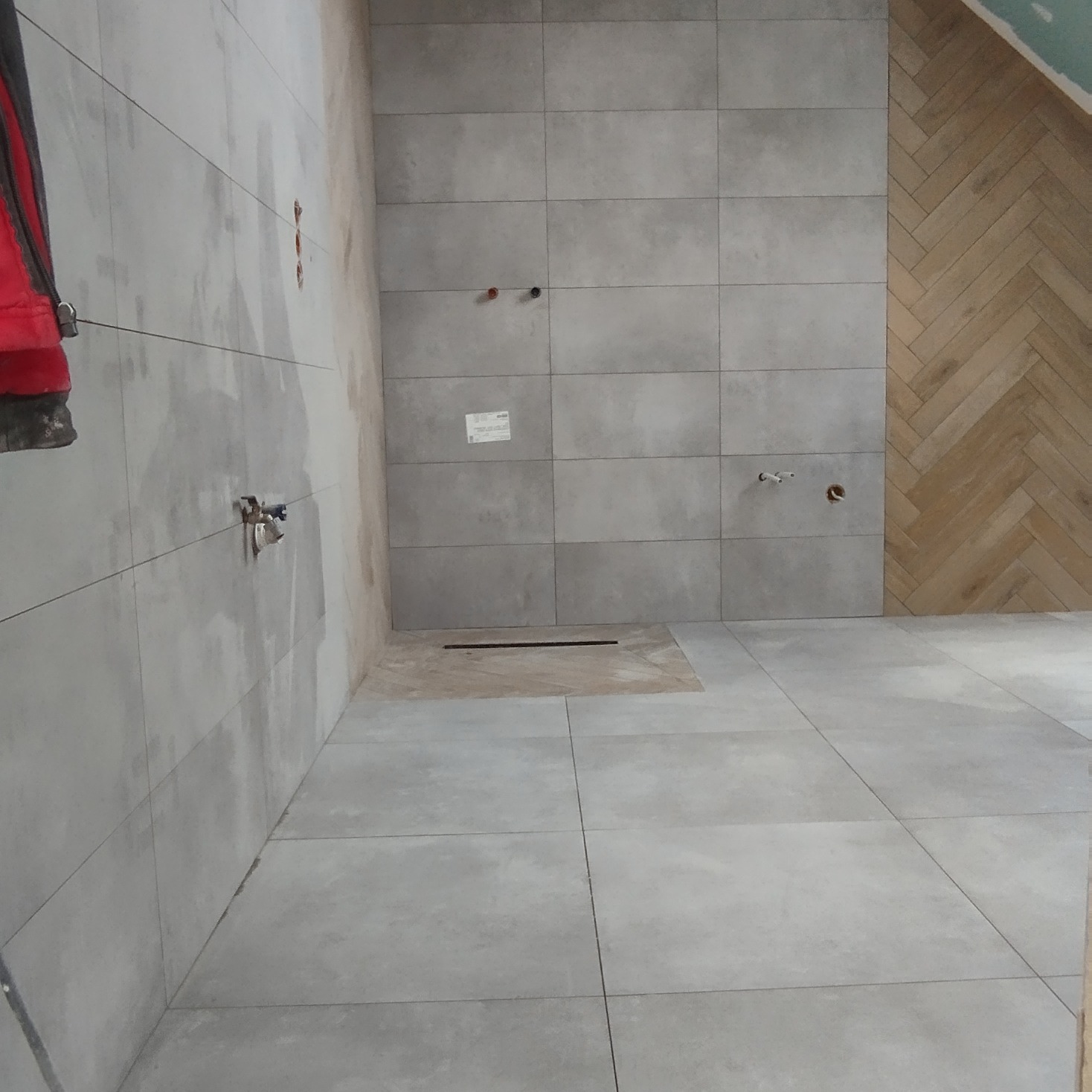 Szare płytki na ścianach i podłodze łazienki w trakcie remontu, widoczny odpływ liniowy i fragment ściany wyłożonej drewnopodobną płytką w jodełkę, z niedokończonymi fugami przy podłodze.