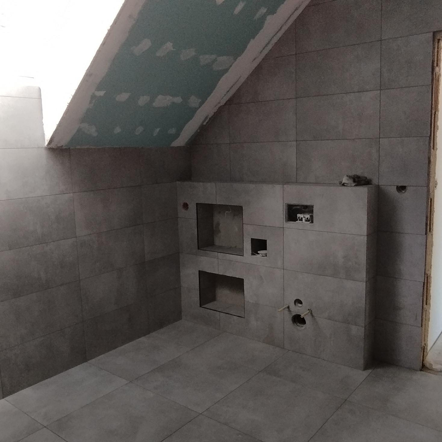 Szara łazienka w trakcie remontu na poddaszu ze skośnym sufitem, widoczne wnęki w ścianie, przygotowane otwory na instalacje i położone płytki podłogowe i ścienne.