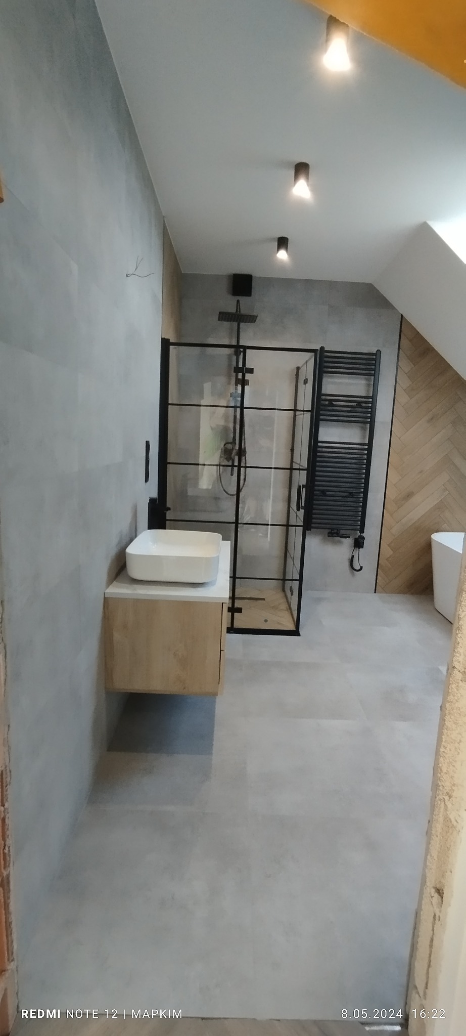 Nowoczesna łazienka z kabiną prysznicową typu walk-in z czarnymi ramami, wisząca szafka z umywalką nablatową, grzejnik drabinkowy w czarnym kolorze oraz oświetlenie punktowe.