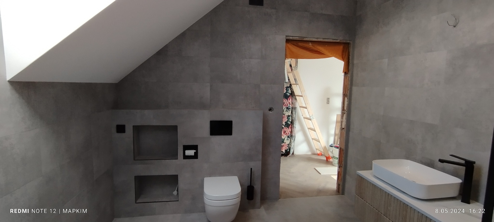 Wykończona łazienka na poddaszu ze skośnym sufitem, szare płytki imitujące beton, wisząca toaleta, umywalka nablatowa i widok na remontowane pomieszczenie z drabiną.