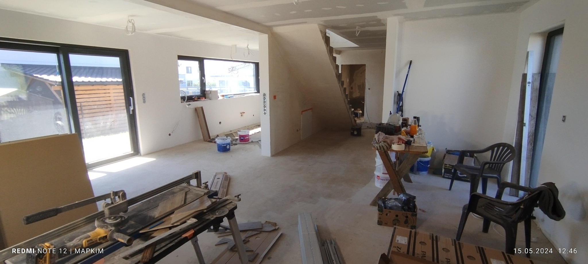 Wnętrze w trakcie remontu: surowe ściany, narzędzia do cięcia płytek, farby, widok na schody i okno z widokiem na zewnątrz.