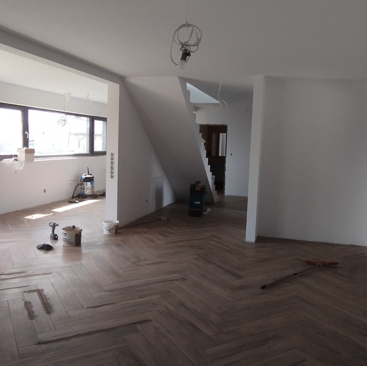 Wnętrze w trakcie remontu: parkiet ułożony w jodełkę, ślady po lakierowaniu, narzędzia (wiertarka, pędzle, wiadra) i odkurzacz budowlany widoczne w przestrzeni z surowymi ścianami i instalacją...