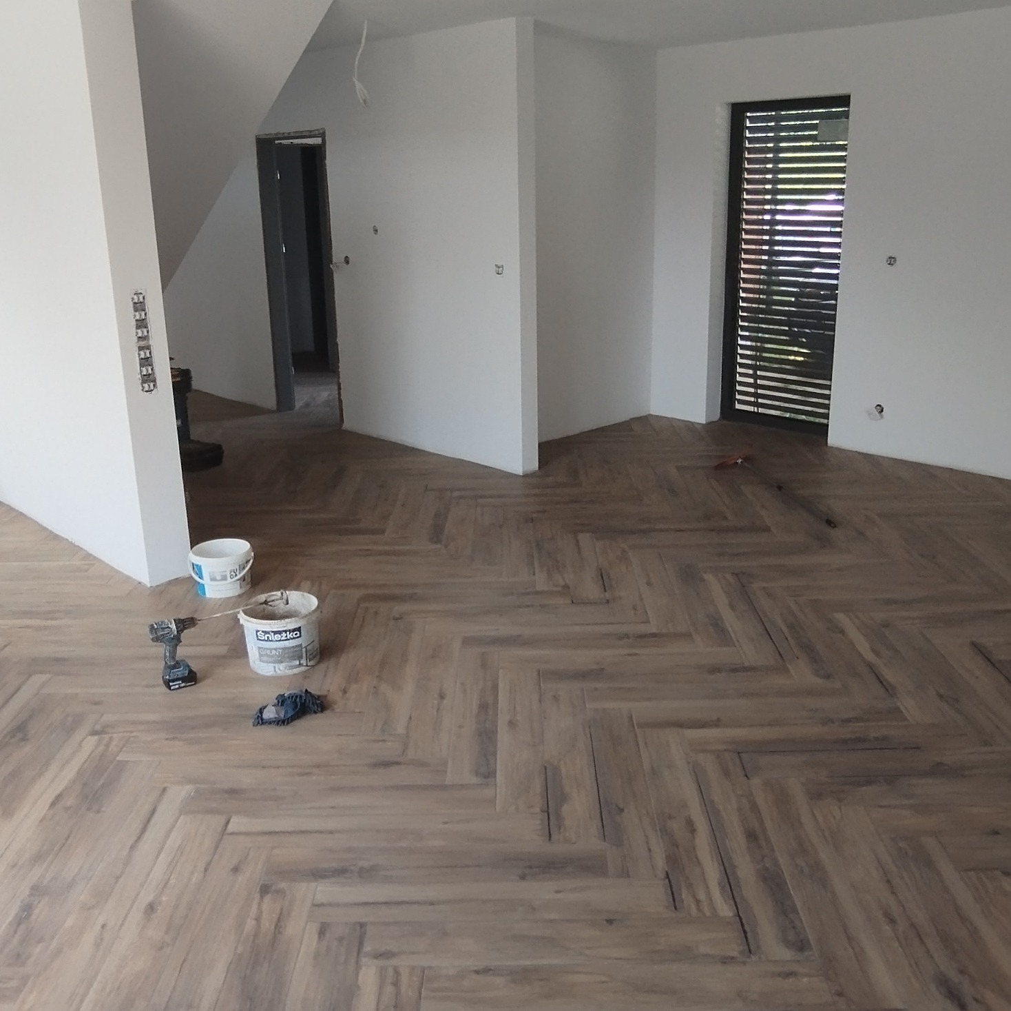 Ułożona podłoga z paneli w jodełkę w pustym, świeżo wyremontowanym pokoju z białymi ścianami; widoczne wiadro z gruntem Śnieżka, wkrętarka akumulatorowa i rękawice robocze.