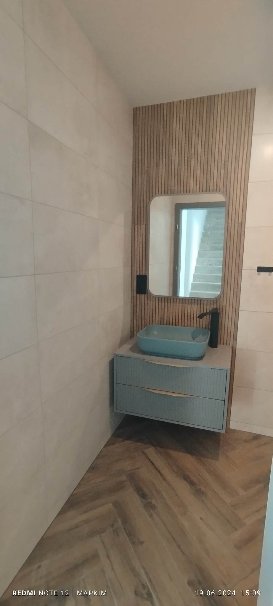 Nowoczesna łazienka z umywalką w kolorze turkusowym, szafką z dwoma szufladami i lustrem na tle ściany z drewnianych listewek, parkiet ułożony w jodełkę.