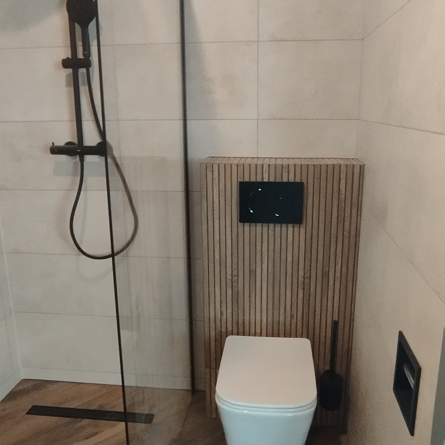 Nowoczesna łazienka z kabiną prysznicową typu walk-in z czarną armaturą, podwieszana toaleta z panelem sterującym na obudowie z drewnianych listewek.