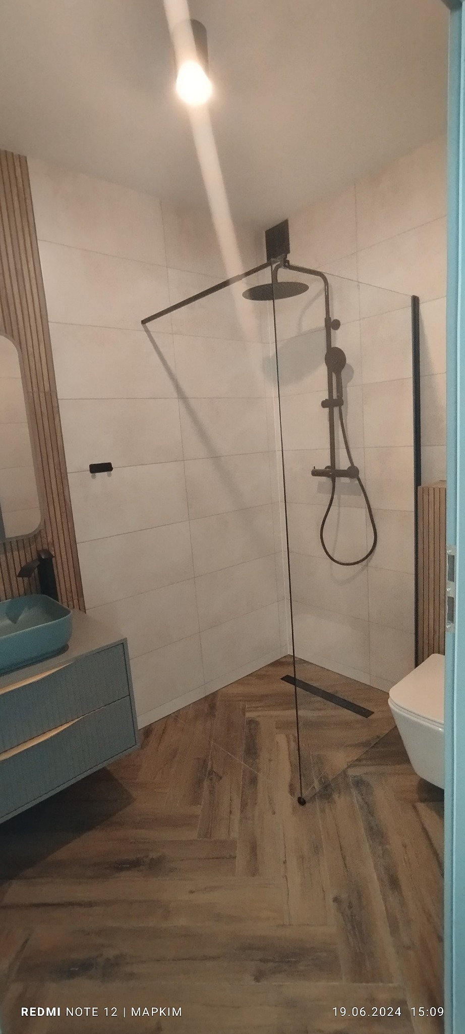 Nowoczesna łazienka z kabiną prysznicową typu walk-in z czarnym osprzętem, umywalką w kolorze turkusowym i drewnianą okładziną ściany.