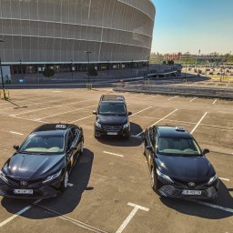 Partner Taxi - Trzy czarne taksówki (dwa sedany Toyota Camry i jeden van Mercedes-Benz) ustawione na parkingu przed stadionem miejskim w słoneczny dzień.
