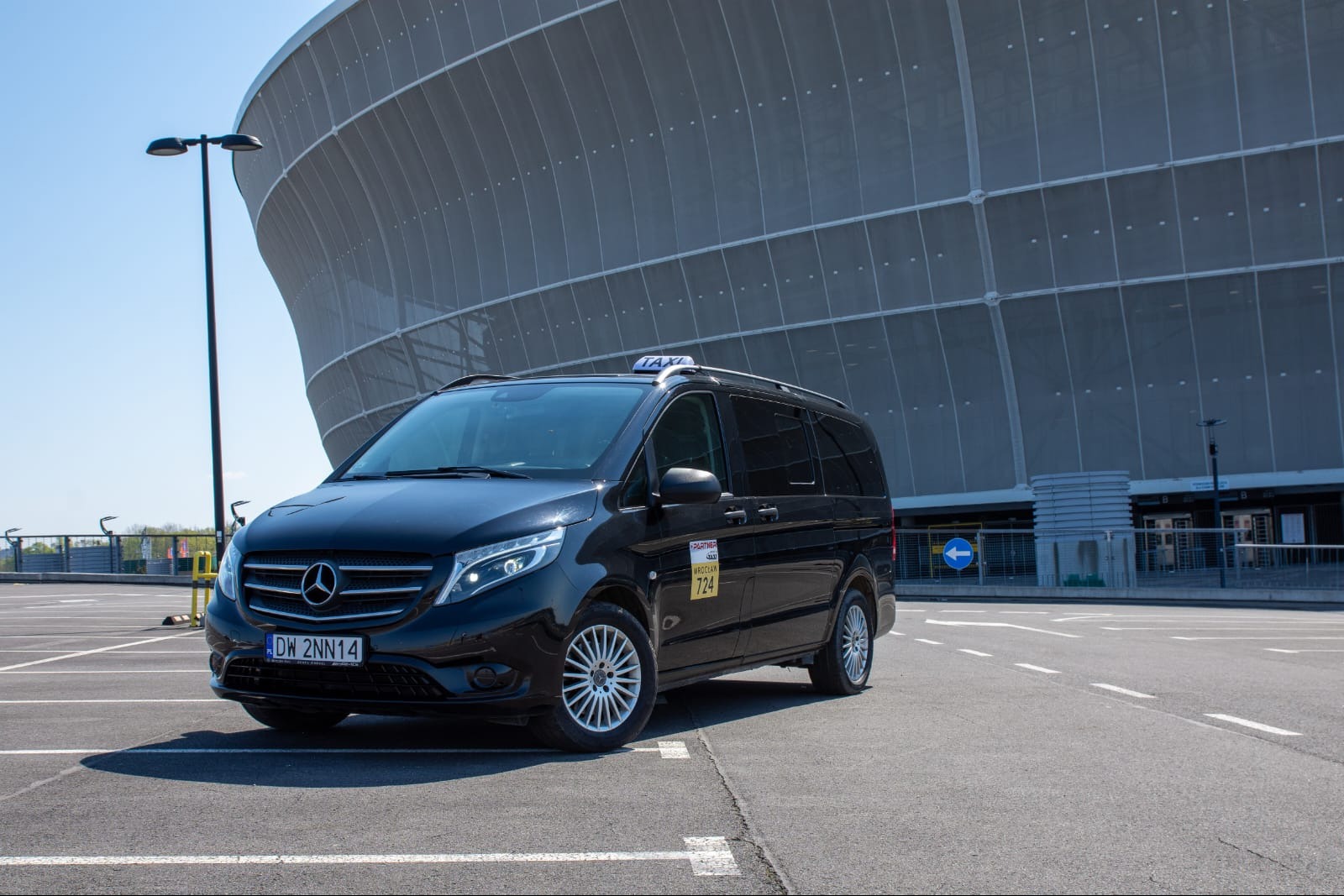Czarny Mercedes-Benz Vito Taxi z tabliczką 'TAXI' na dachu, zaparkowany na parkingu przed nowoczesnym stadionem. Na boku pojazdu widoczna naklejka z numerem telefonu i napisem 'Partner Taxi Wrocław'.