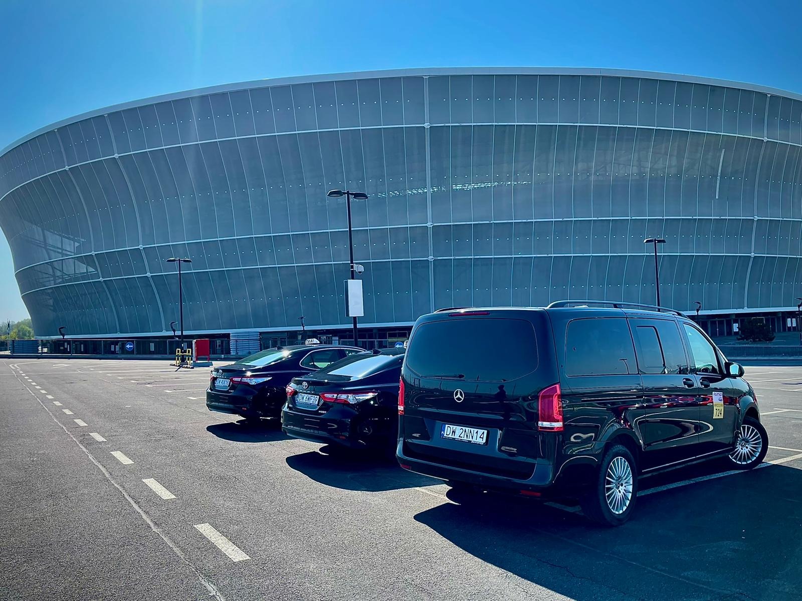 Trzy czarne samochody, w tym van Mercedes z numerem bocznym taxi 724, zaparkowane na parkingu przed nowoczesnym stadionem w słoneczny dzień.