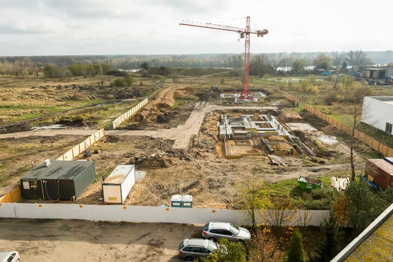 Wysoki kąt widzenia na plac budowy z fundamentami, dźwigiem w tle, barakami i zaparkowanymi samochodami na obrzeżach.
