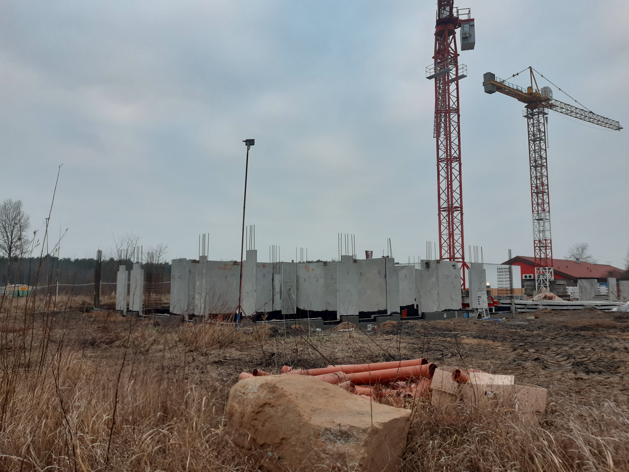 Wznoszenie ścian budynku z prefabrykatów betonowych na placu budowy z dwoma żurawiami w tle, widoczne rury i gruz.