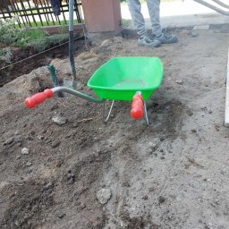 Przemyslaw Szota - Zielona taczka dziecięca z czerwonymi rączkami na ziemi, obok wbita łopata i osoba w szarych spodniach z narzędziem w dłoniach w tle, teren przygotowany pod prace ziemne.