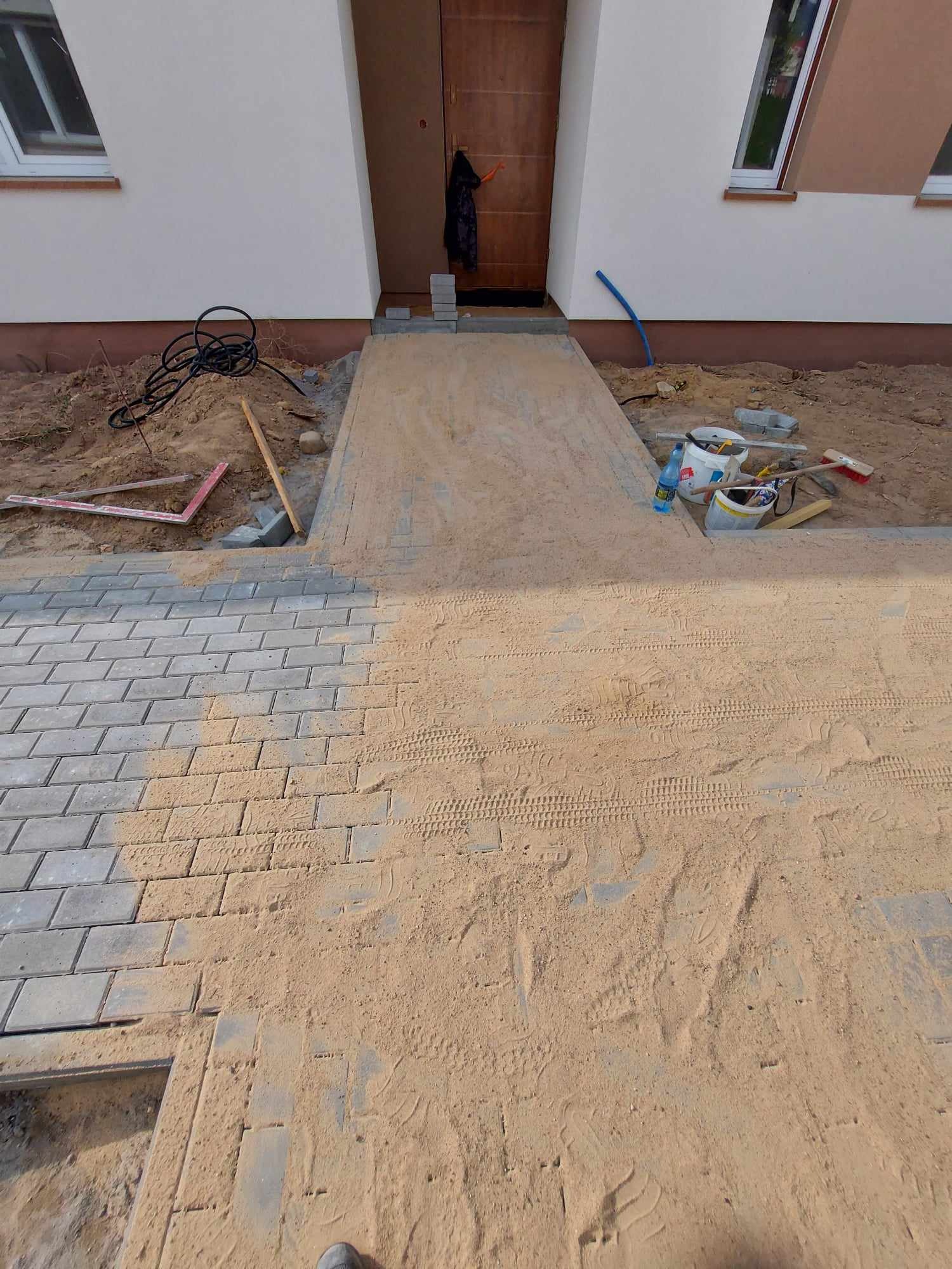 Fragment chodnika z szarej kostki brukowej w trakcie układania, widoczne ślady opon i narzędzia budowlane na piasku przed wejściem do domu.