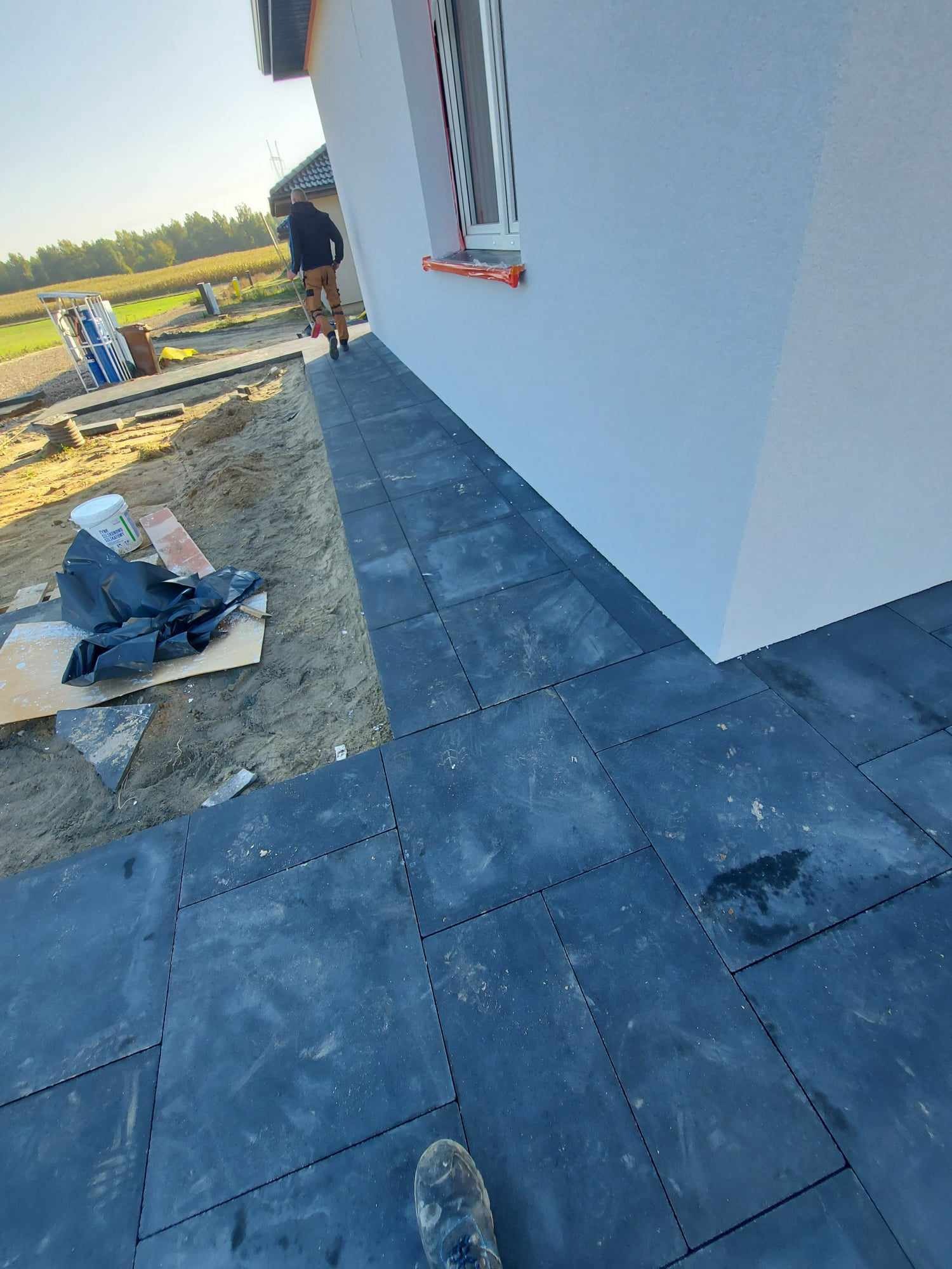 Świeżo ułożona, ciemnoszara kostka brukowa przylegająca do białej ściany budynku, z widocznym przygotowanym podłożem i pracownikiem w oddali.