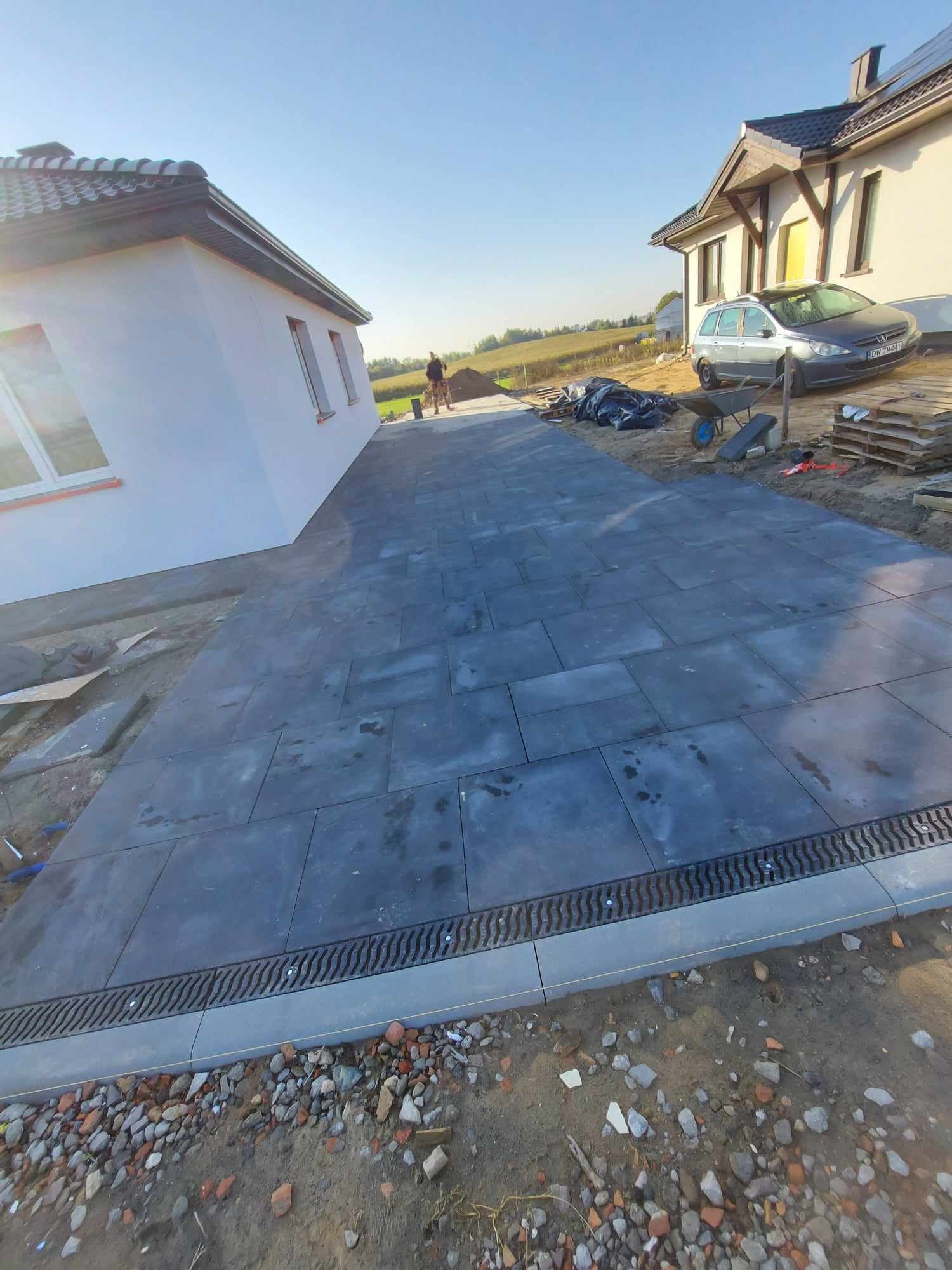 Ułożona, szara kostka brukowa na podjeździe z widocznym odpływem liniowym, w tle domy i osoba pracująca.