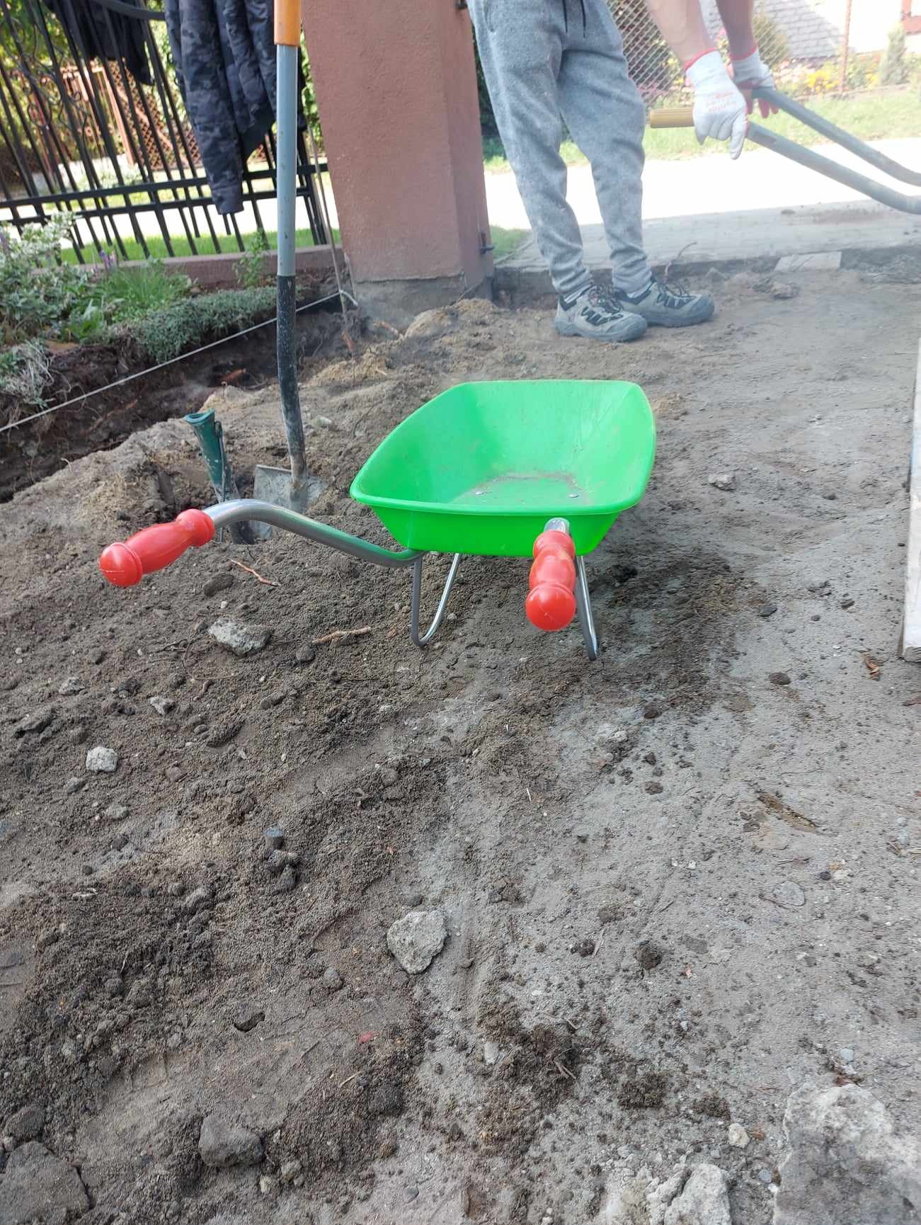 Zielona taczka dziecięca z czerwonymi rączkami na ziemi, obok wbita łopata i osoba w szarych spodniach z narzędziem w dłoniach w tle, teren przygotowany pod prace ziemne.