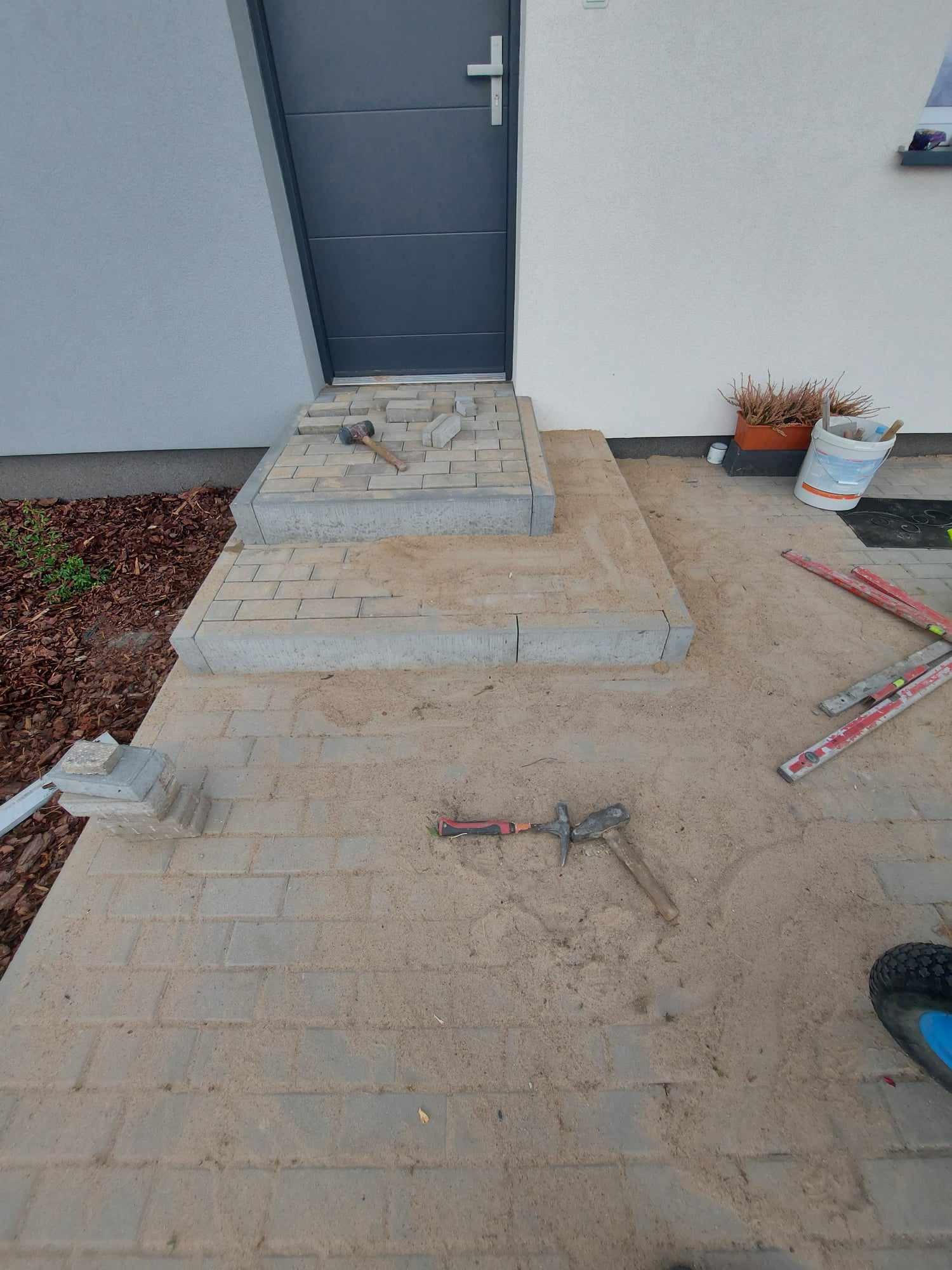 Budowa schodów wejściowych z kostki brukowej: widoczne narzędzia, poziomice i piasek na powierzchni kostki, częściowo ułożone stopnie.