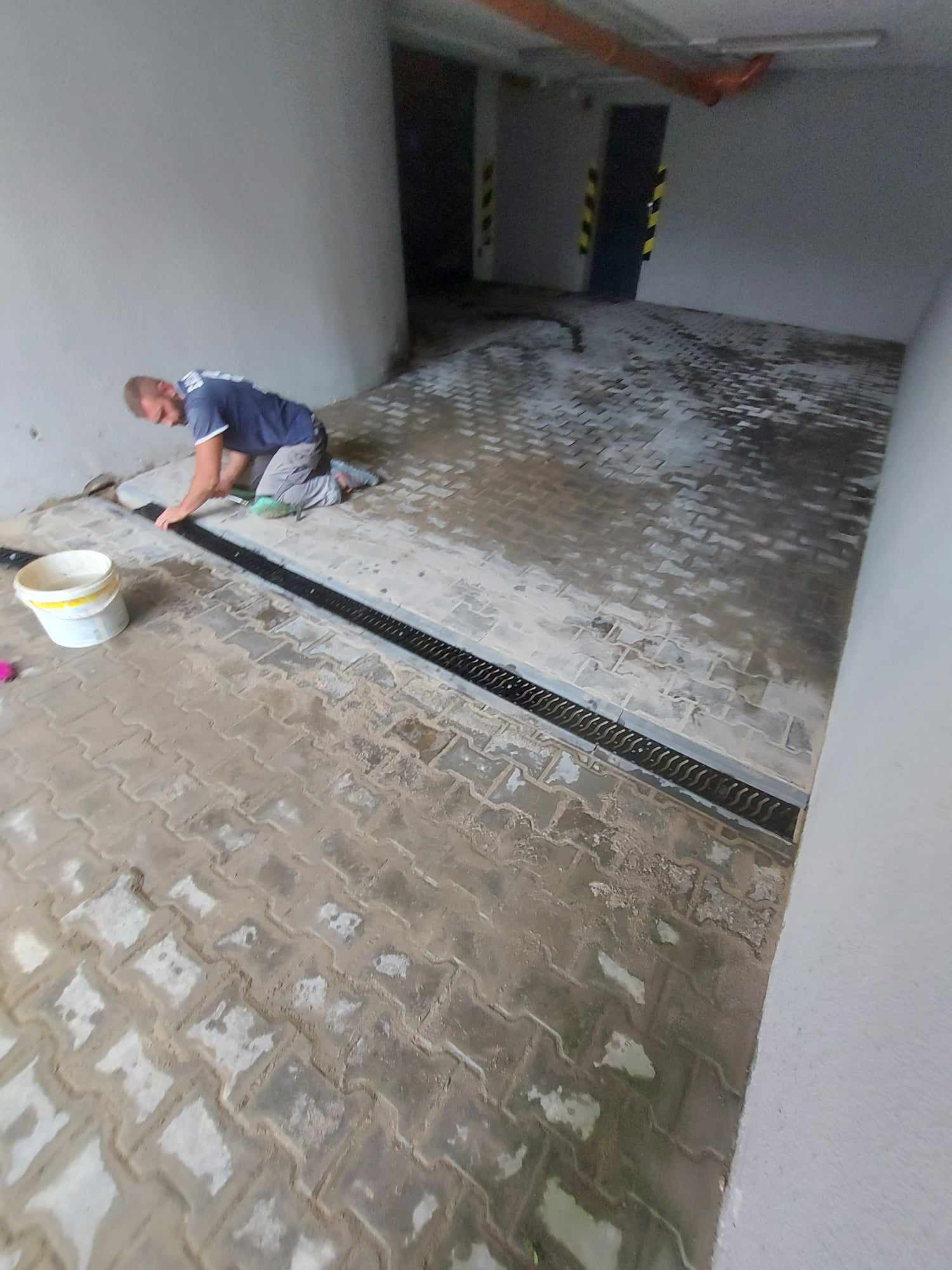 Pracownik na kolanach wyrównuje czarną kratkę odpływową na świeżo ułożonej, mokrej kostce brukowej w garażu podziemnym. Widoczny biały kubeł i narzędzia.