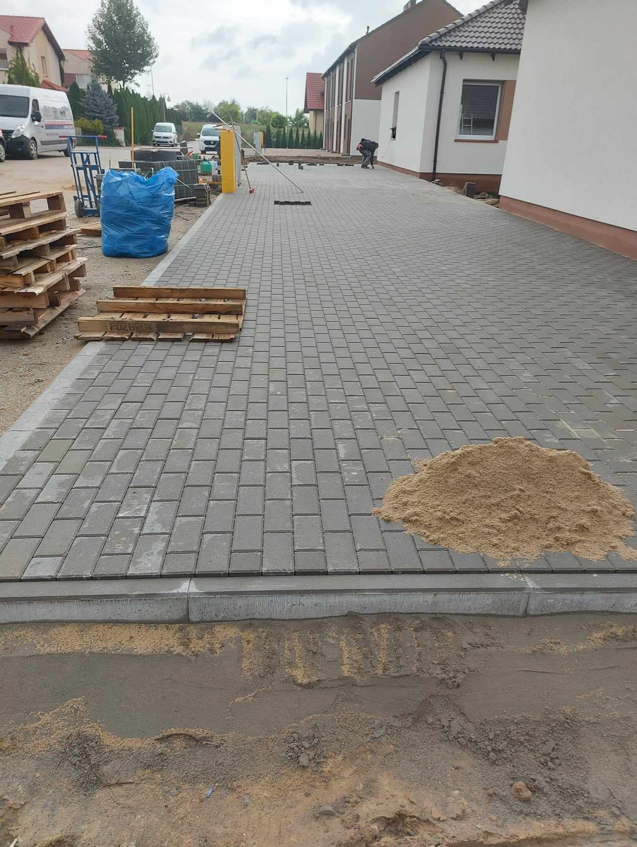 Układanie szarej kostki brukowej na posesji, widoczny stos kostki, piasek i pracownik w oddali.