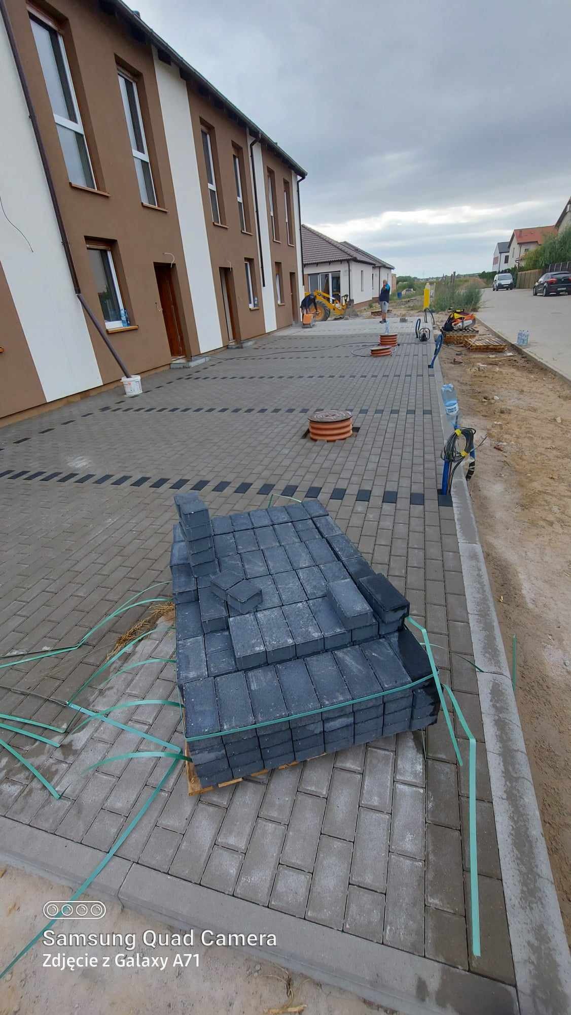 Stos szarych kostek brukowych na palecie, częściowo ułożona nawierzchnia z ciemnymi akcentami, w tle budynek mieszkalny i prace ziemne z użyciem minikoparki.