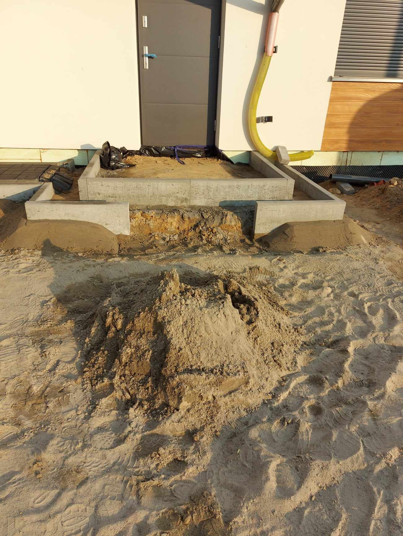 Przygotowanie terenu pod schody wejściowe do domu z widoczną warstwą piasku i betonowymi obrzeżami, rura spustowa na elewacji budynku.