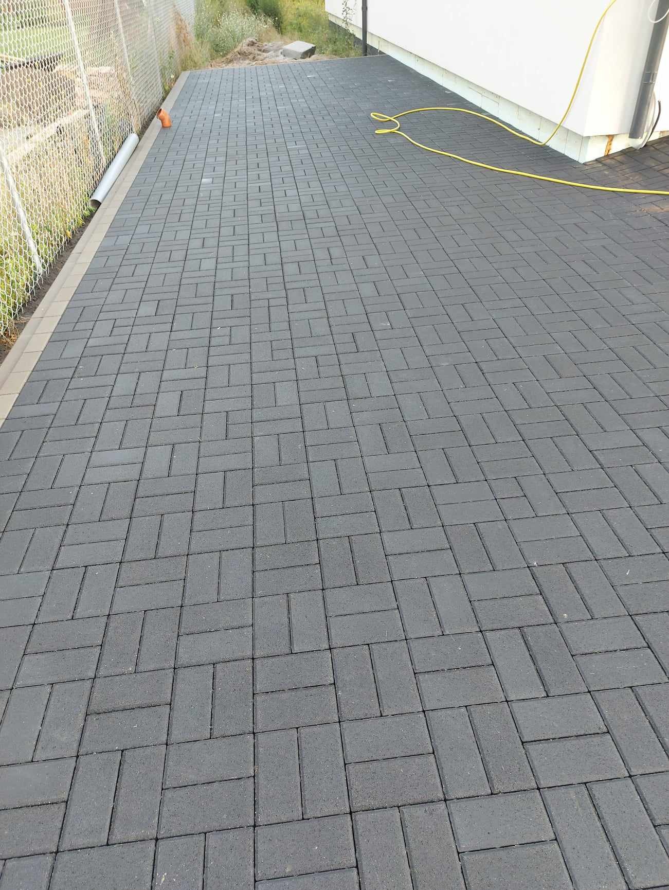 Nowo ułożona, grafitowa kostka brukowa w wzór jodełki na posesji, widoczny fragment ogrodzenia z siatki i żółty wąż ogrodowy przy białej ścianie budynku.