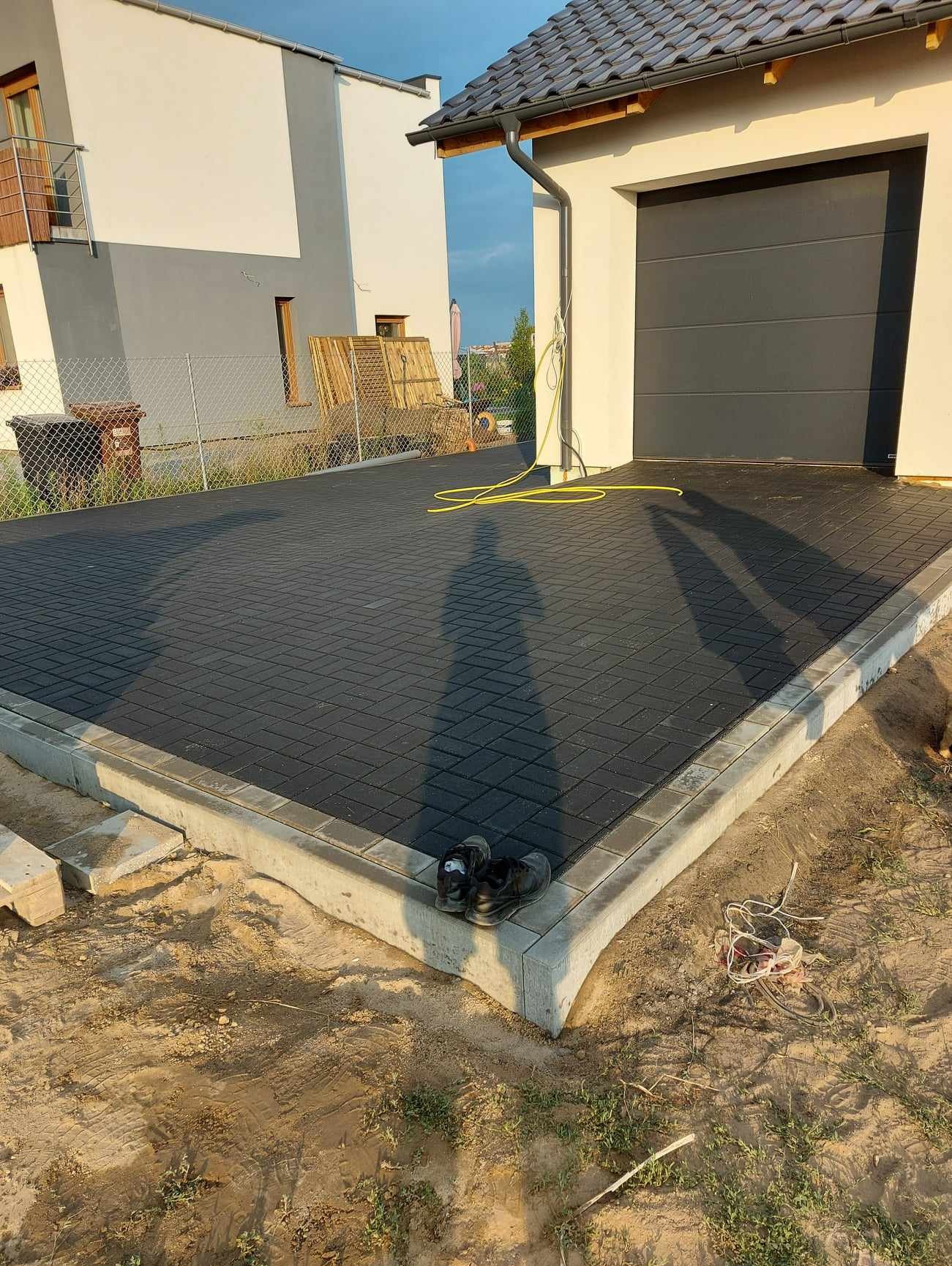 Ułożona, ciemnoszara kostka brukowa na podjeździe do garażu, z widocznym cieniem osoby fotografującej, para roboczych butów stoi na obrzeżu. Żółty wąż ogrodowy leży na kostce, a w tle widoczny dom...