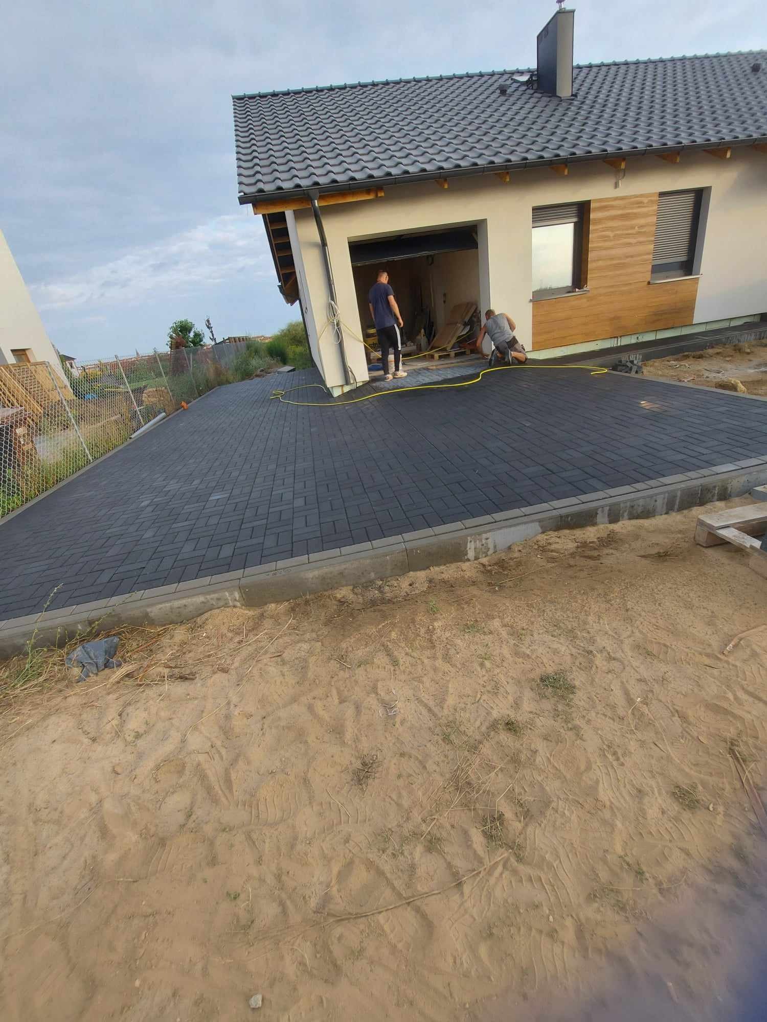 Ciemnoszara kostka brukowa ułożona przed nowoczesnym domem z garażem, dwóch pracowników na kolanach przy garażu, widoczny przewód żółty, piasek na pierwszym planie.