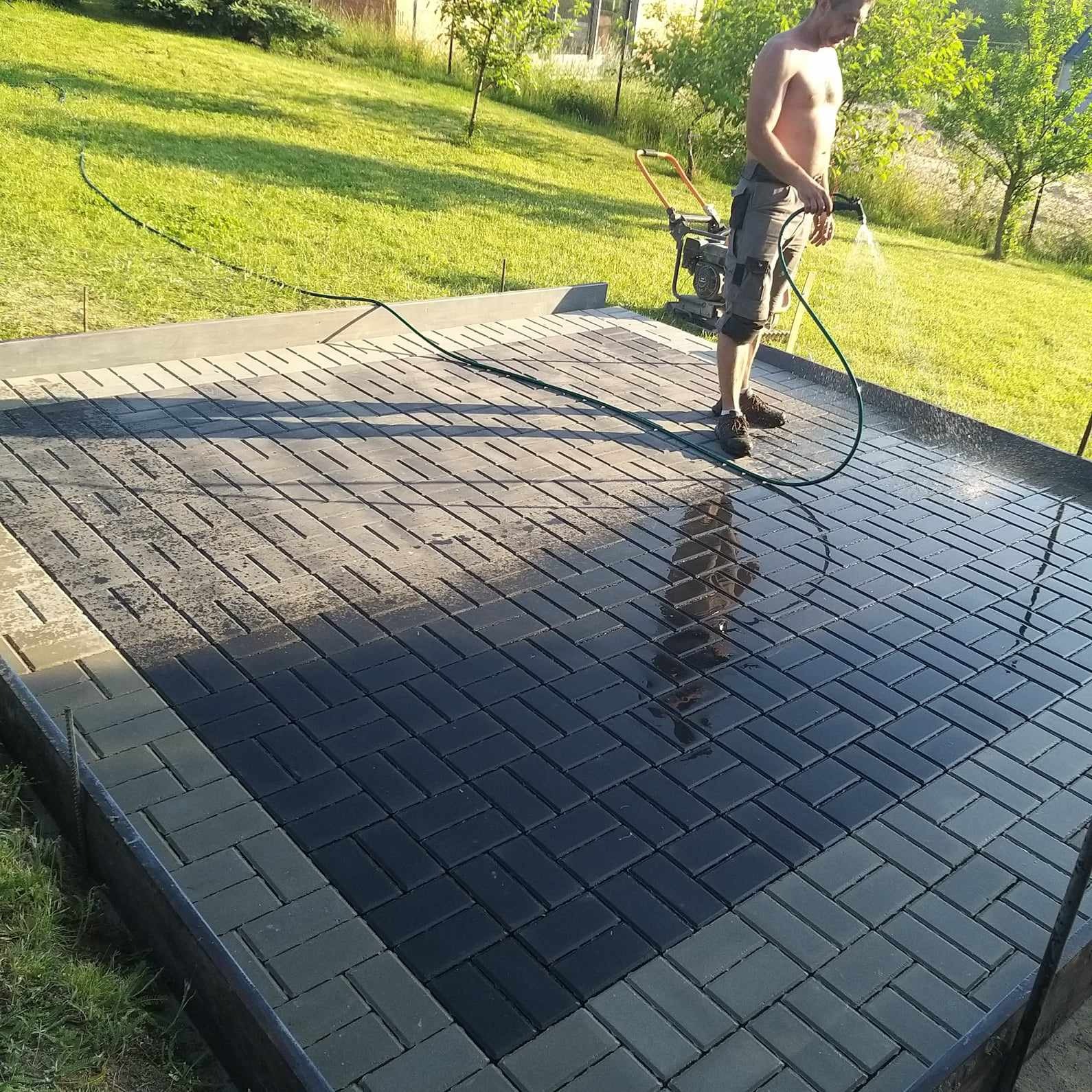 Mężczyzna w Poznaniu spłukuje świeżo ułożoną, ciemnoszarą kostkę brukową w jodełkę, z widoczną maszyną do zagęszczania w tle.