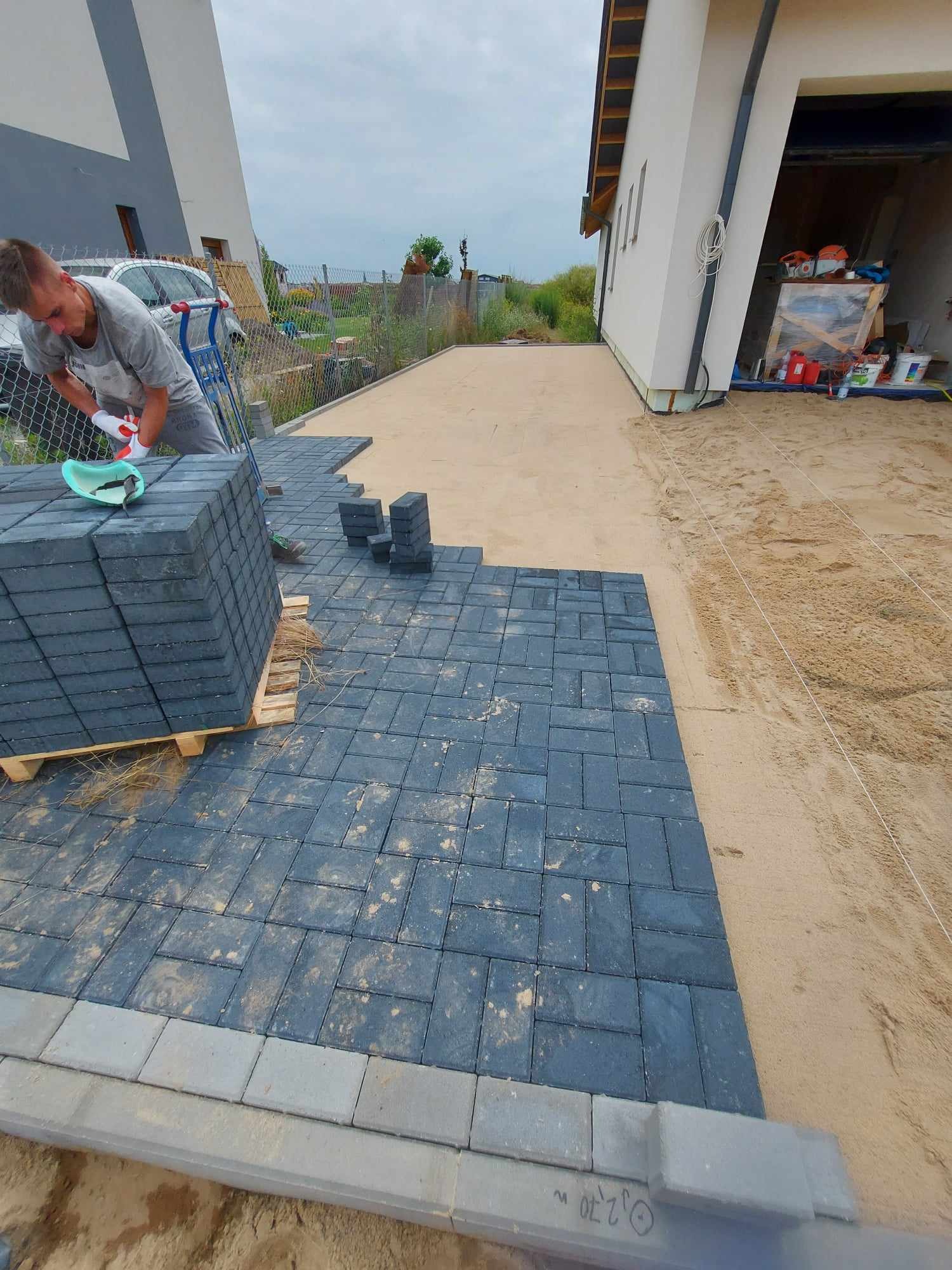 Pracownik w rękawicach roboczych układa ciemnoszarą kostkę brukową na podsypce piaskowej, obok paleta z kostką, w tle dom jednorodzinny i ogrodzenie z siatki.