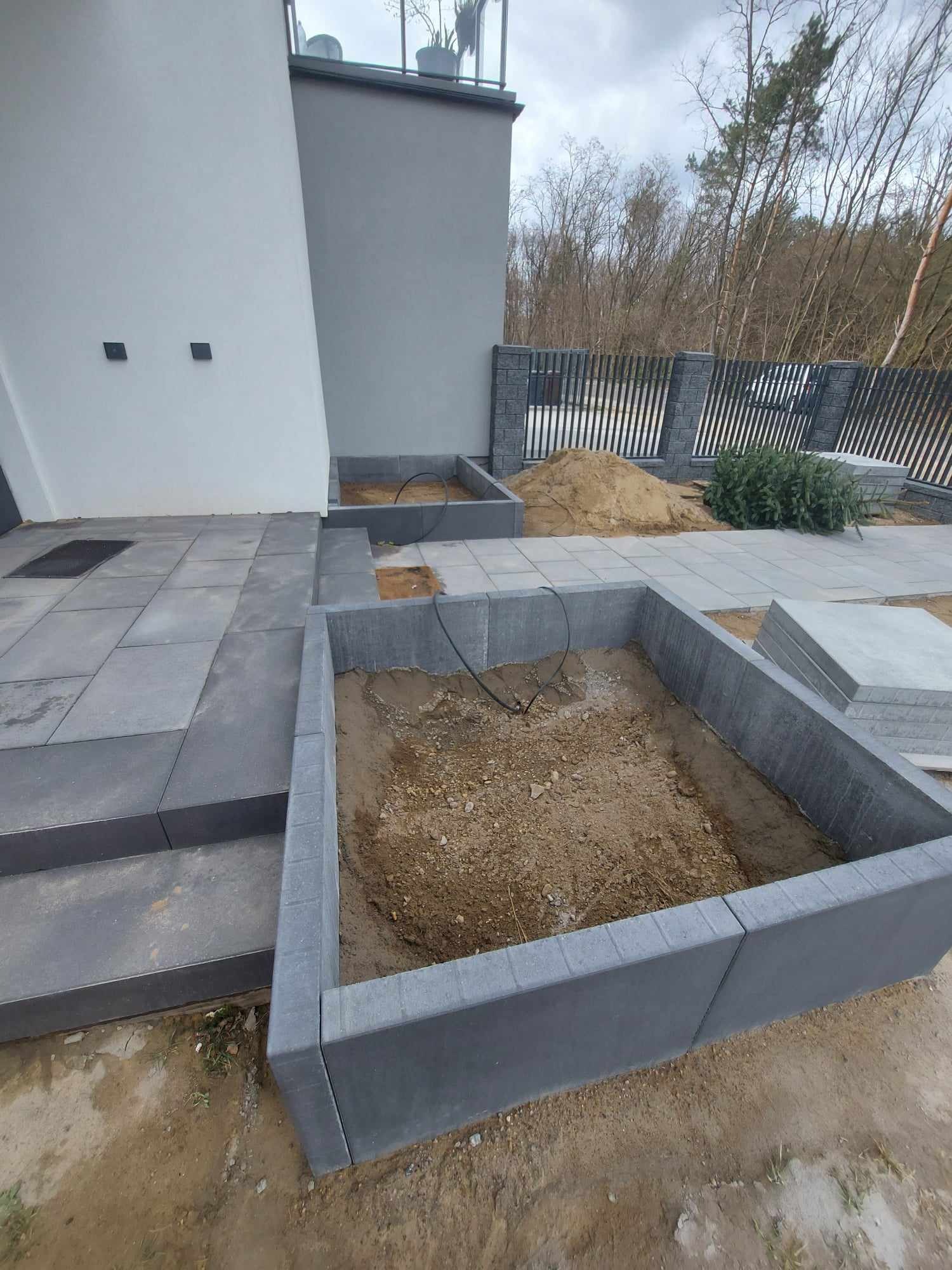 Przygotowane rabaty kwiatowe z szarej kostki brukowej, częściowo ułożony chodnik z kostki, ogrodzenie panelowe w tle, widok na dom i drzewa.