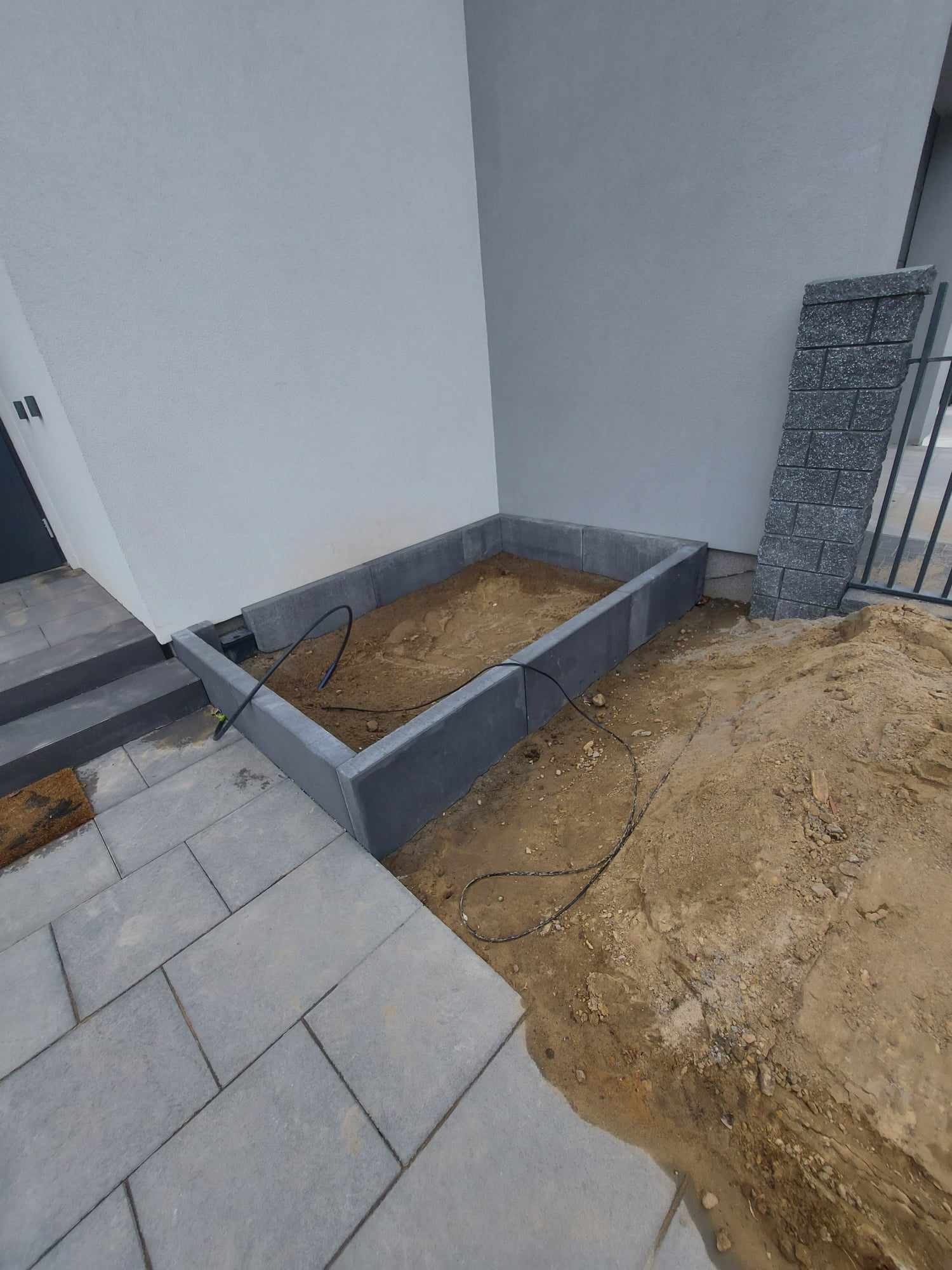 Przygotowany fundament z obrzeżami z szarej kostki betonowej na rabatę kwiatową przy ścianie budynku, widoczny kabel doprowadzony do wnętrza rabaty.