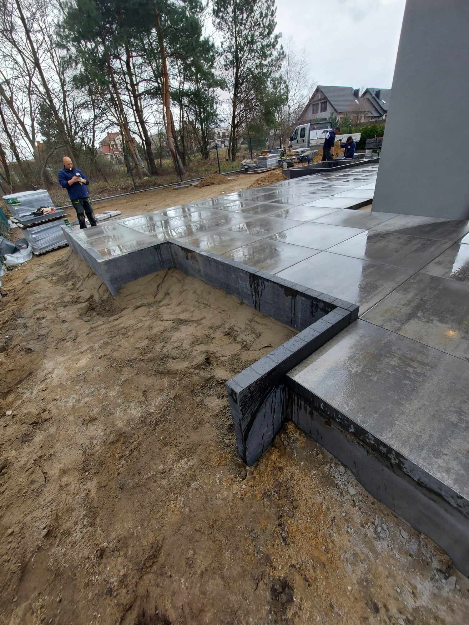 Świeżo ułożona, mokra nawierzchnia z dużych, szarych płyt betonowych, z widocznym fragmentem obrzeża i przygotowanym podłożem z piasku. W tle pracownicy i zaparkowany bus.