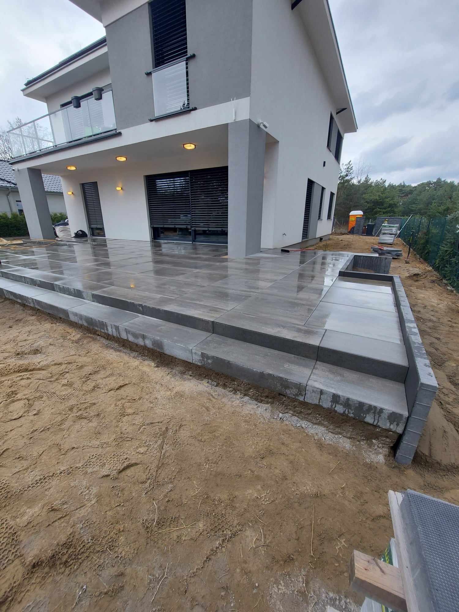 Nowoczesny taras z szarych płyt betonowych, schody z kostki, widok na dwukondygnacyjny dom z balkonem z barierką ze szkła, teren wokół w trakcie prac ziemnych.