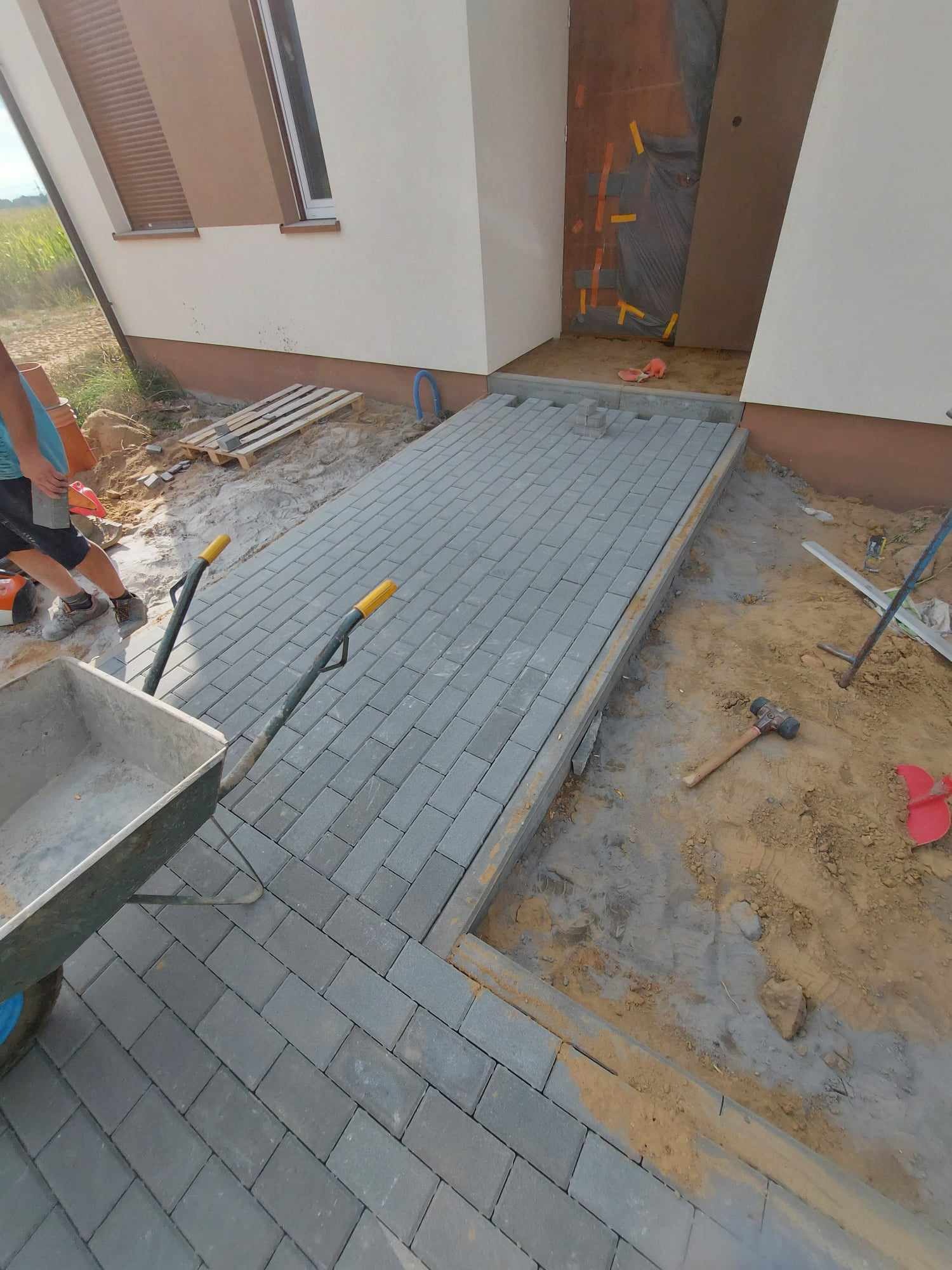 Fragment podjazdu z szarej kostki brukowej w trakcie układania przy nowym domu, widoczny fragment pracownika i taczka z narzędziami.