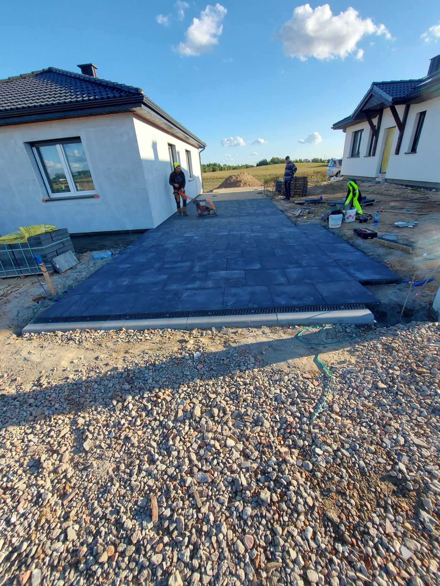 Układanie ciemnoszarej kostki brukowej na podjeździe między dwoma nowymi domami, widoczne krawężniki i drenaż, pracownicy używają zagęszczarki w słoneczny dzień.